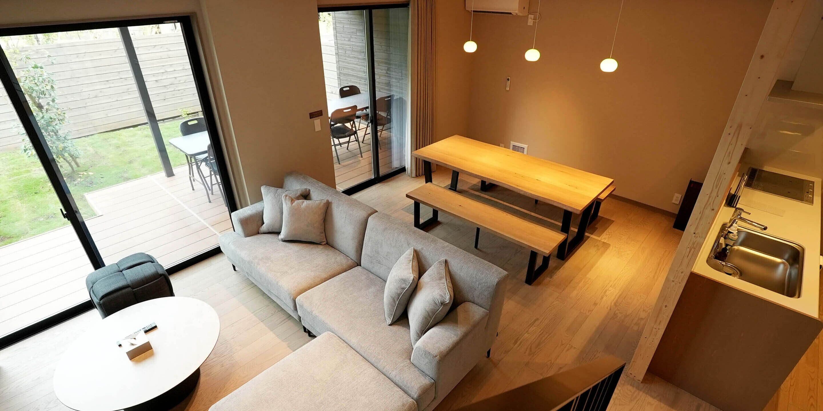 Ein modernes Wohnzimmer mit einer Couch, einem runden Couchtisch und einem Esstisch. Ein großes Schiebe-Glasfenster führt auf eine Terrasse mit Gartenmöbeln.
