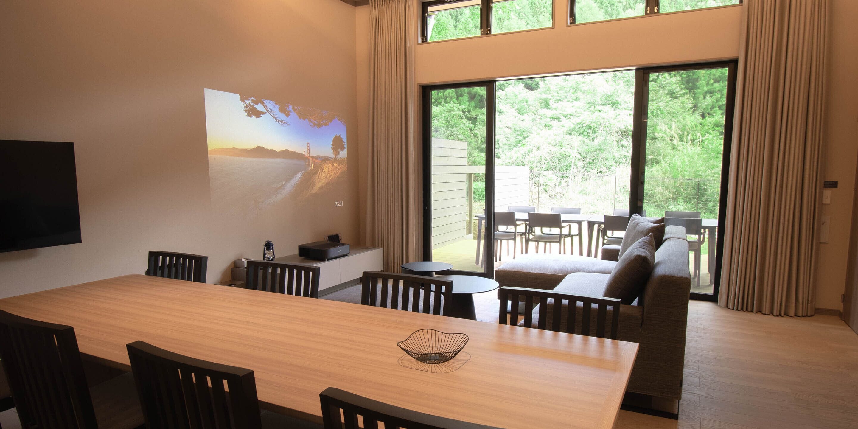 A modern dining area with a wooden table and chairs, a projector screen displaying a scenic image, and a large window offering a view of greenery outside.