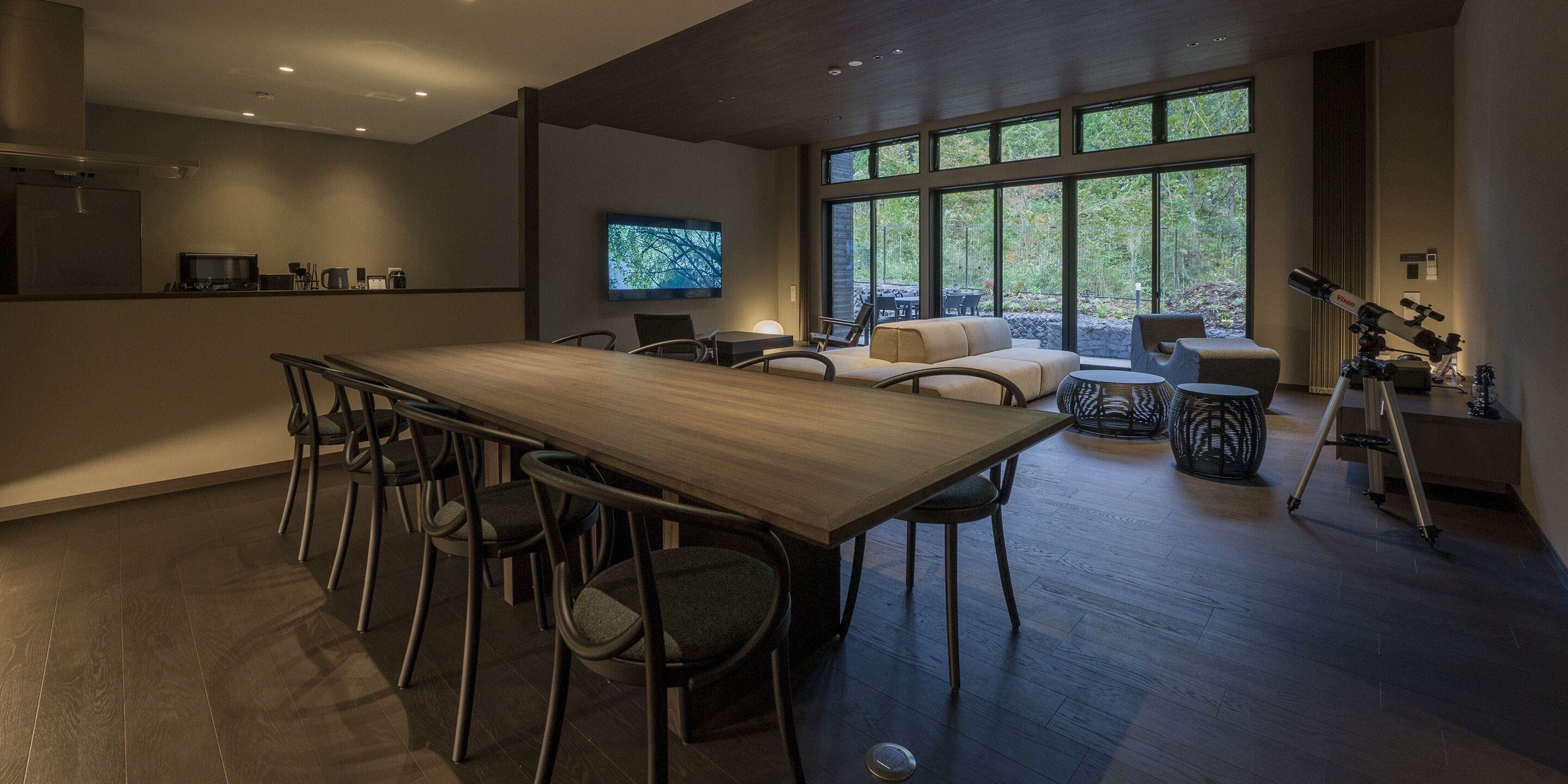 A modern kitchen with a wooden table and chairs, a television, and a large window with a view of trees.