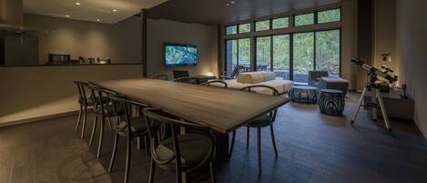 A modern kitchen with a wooden table and chairs, a television, and a large window with a view of trees.