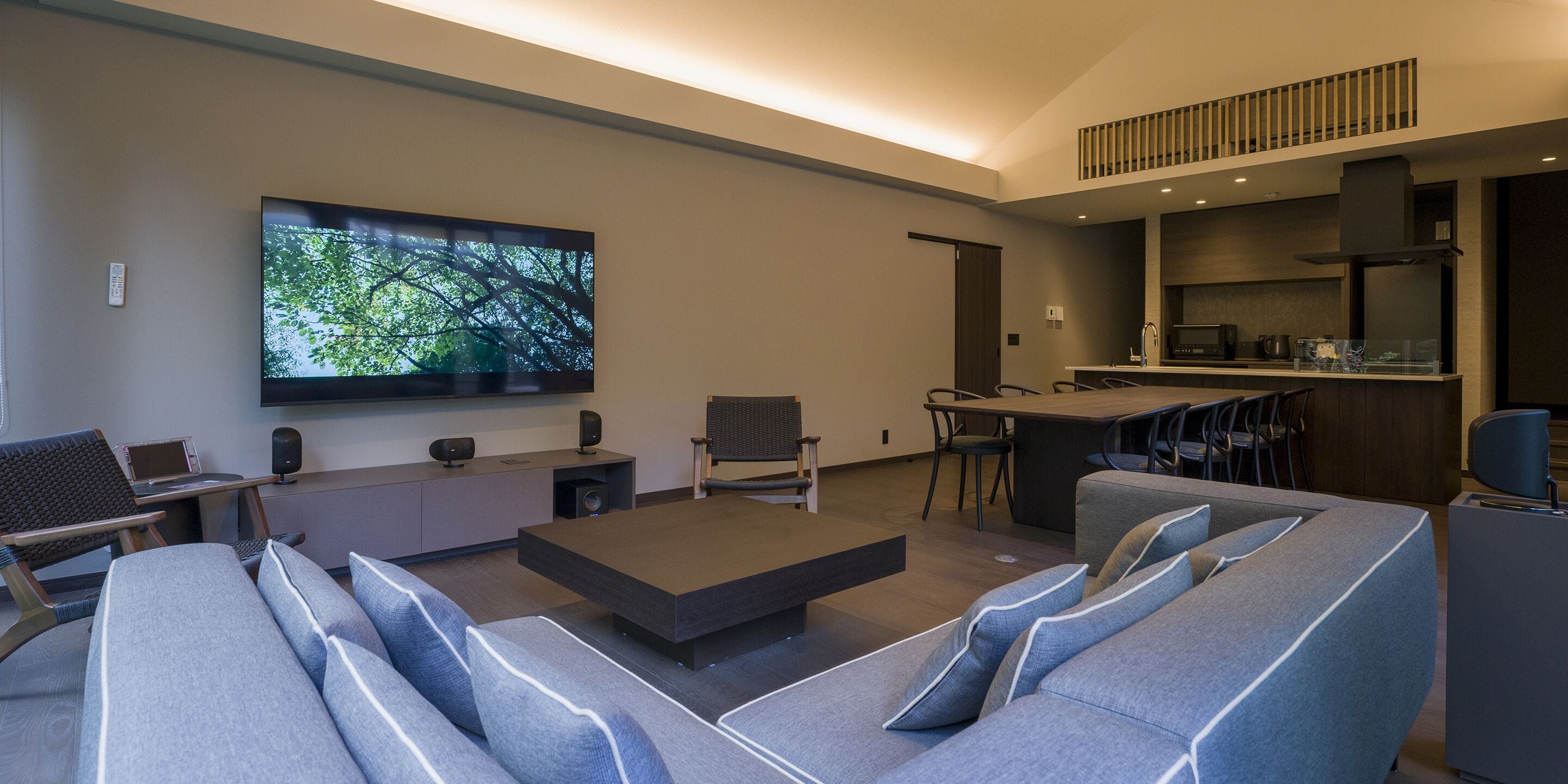 A modern living room with a large flat-screen TV, a sofa, a coffee table, and a kitchen area with a bar.