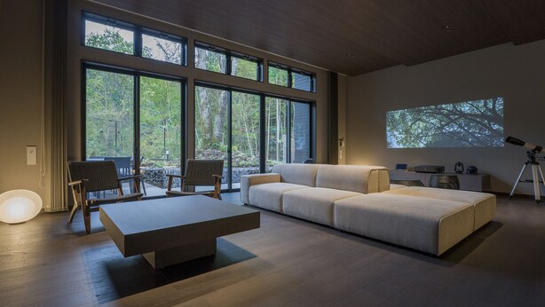 A modern living room with a large sectional sofa, a coffee table, and a dining area with chairs. There is a large window with a view of trees.