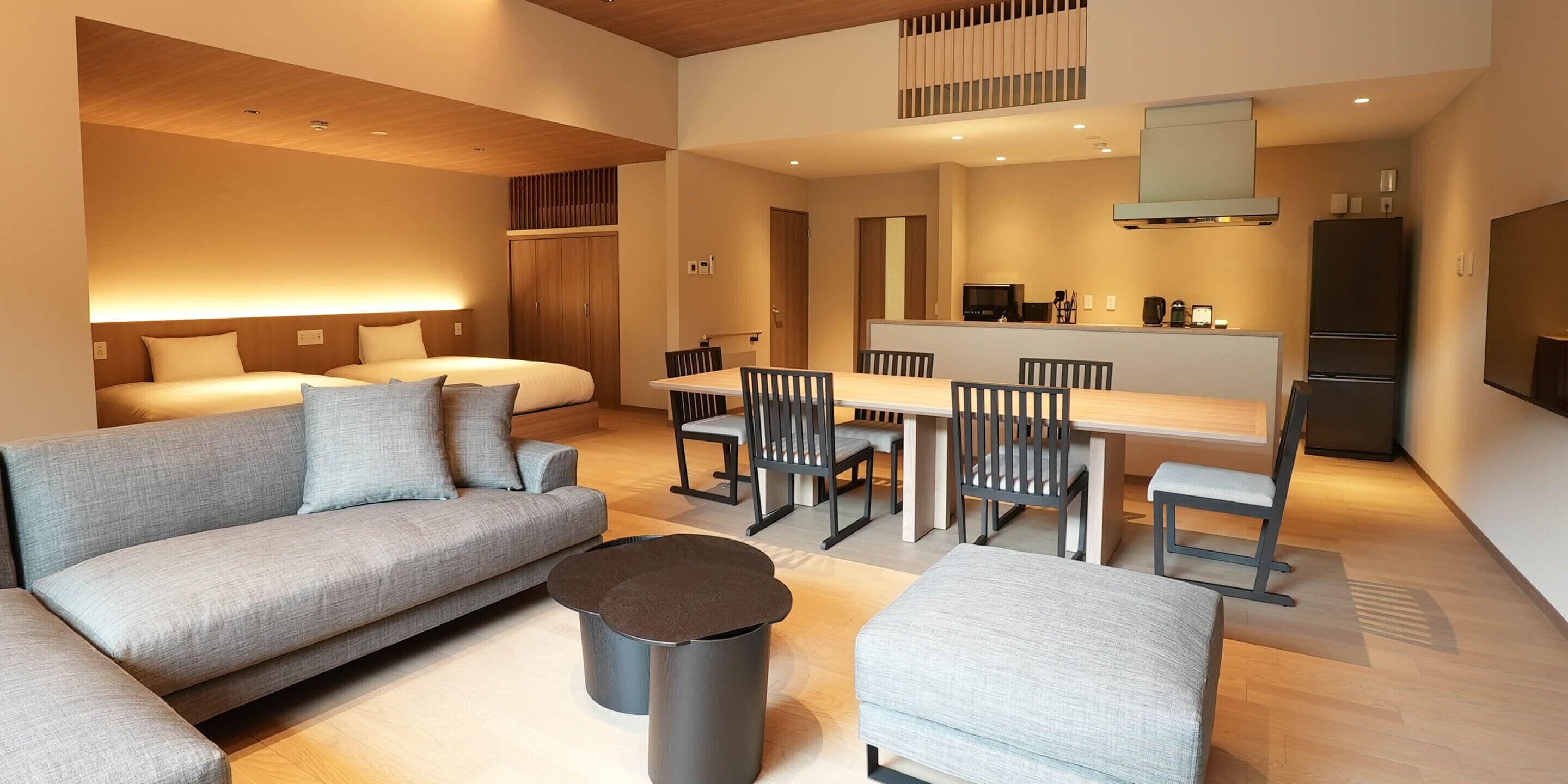 A modern hotel room with a sofa, a dining table with chairs, a kitchen area, and a bed.