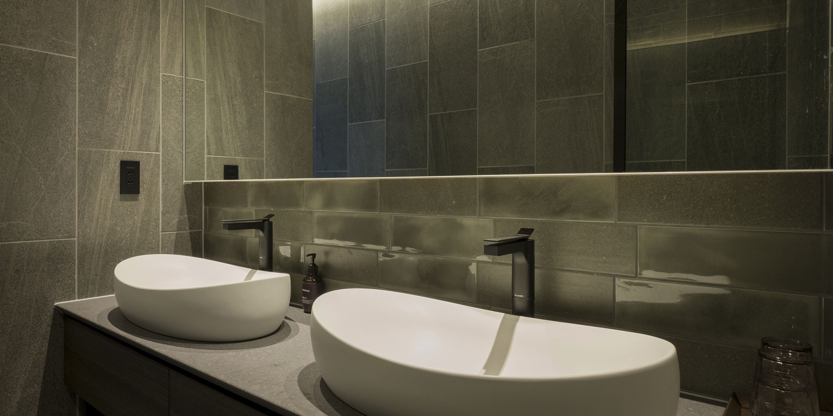 A modern bathroom with two large white sinks, a dark countertop, and a large mirror reflecting the tiled walls.