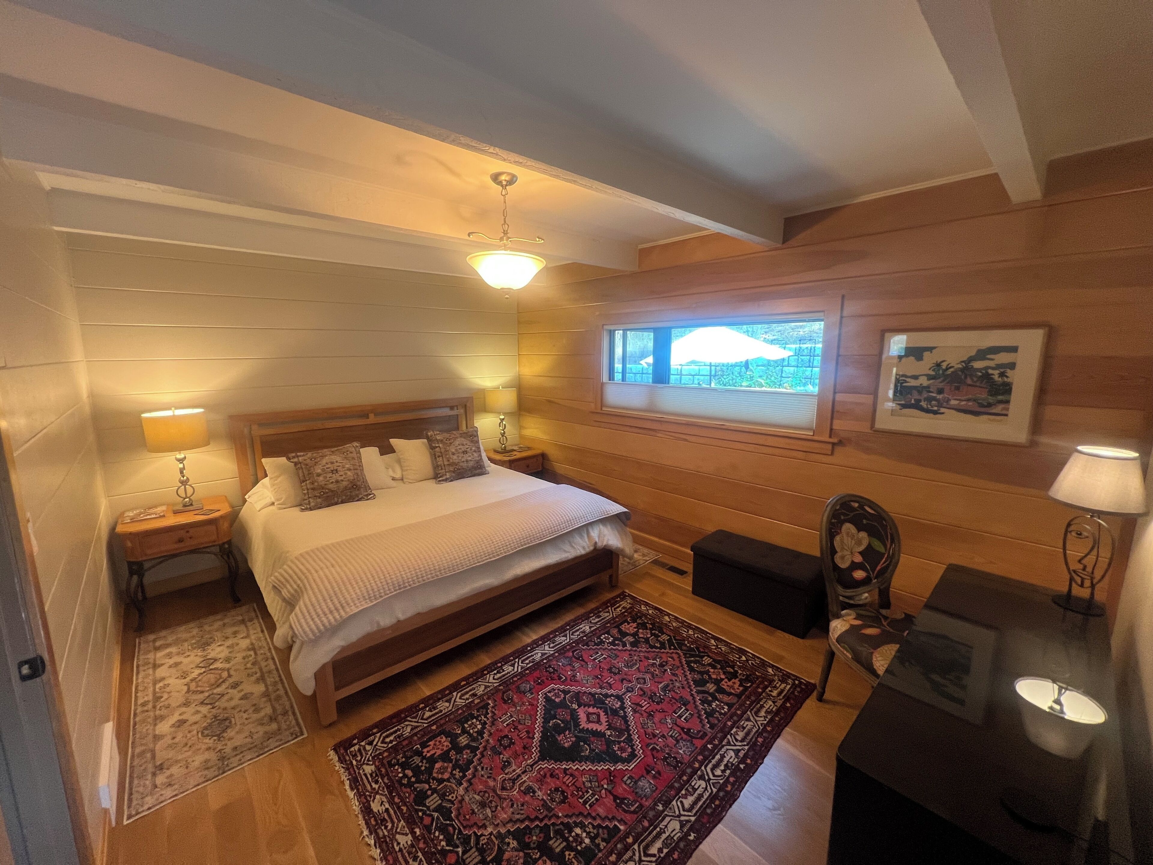Un dormitorio con cabecera de madera, una cama grande, una mesita de noche con lámpara, una ventana con vistas y una pequeña mesa con lámpara.