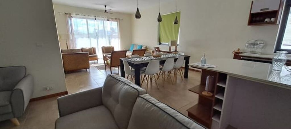 A modern living room with a sofa, dining table, and chairs. There is a kitchen area with a countertop and cabinets.