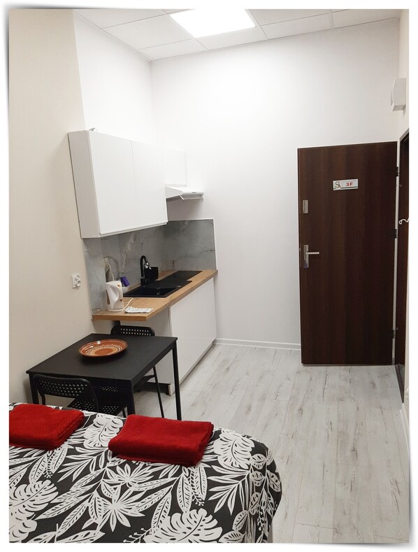 A compact kitchen with a sink, stove, and cabinets, a small dining table with chairs, and a door with a sign.