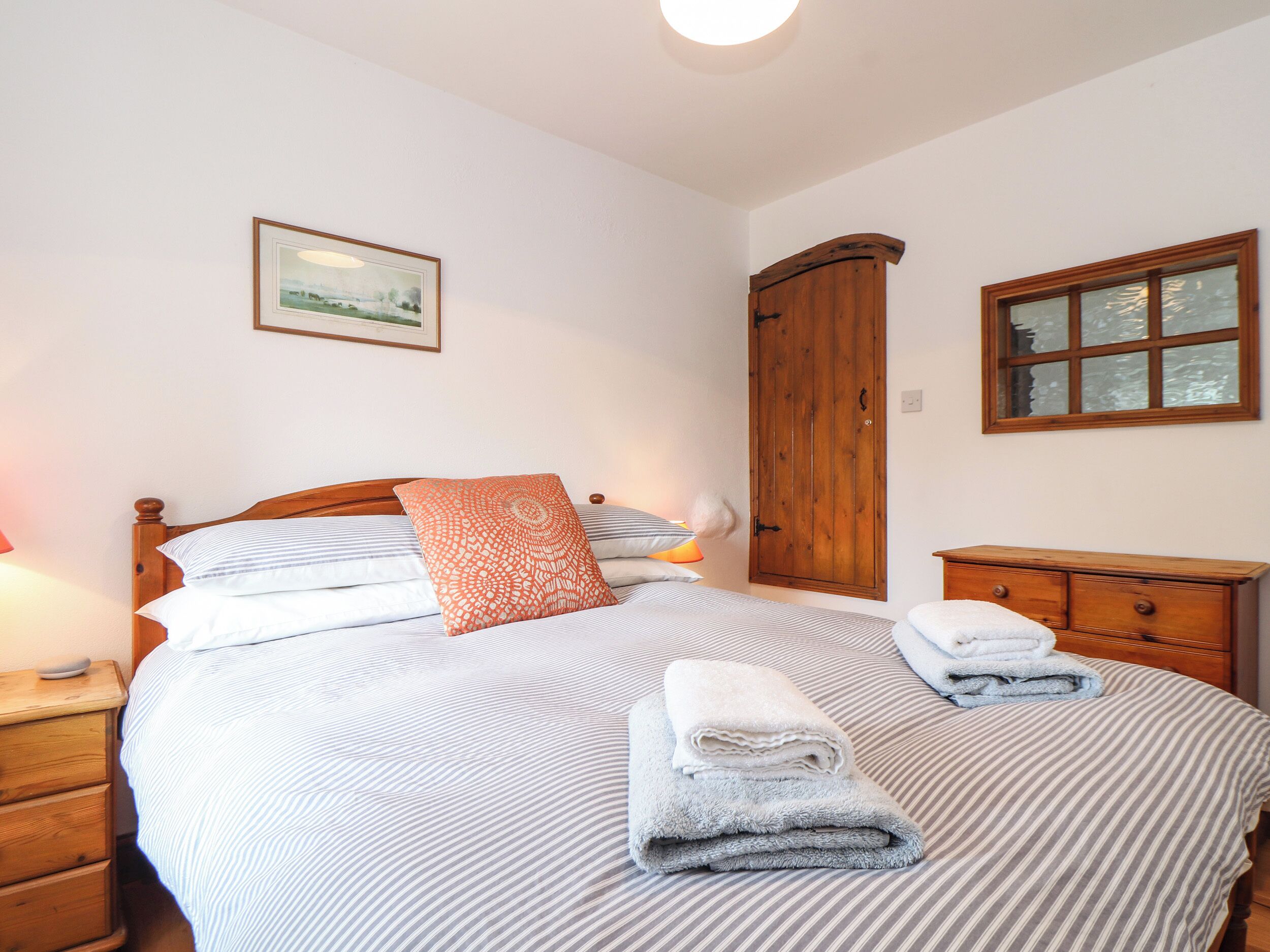 A neatly made bed with striped bedding and a single decorative pillow, a wooden wardrobe, a small bedside table with a lamp, and a framed picture on the wall.