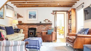 A cozy living room with a fireplace, wooden beams, leather sofas, and a bookshelf. - The Old Barn (Penzance)
