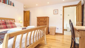 A bedroom with a wooden bed, a wooden dresser, a wooden wardrobe, a bedside table with a lamp, and a shelf with books. - The Old Barn (Penzance)