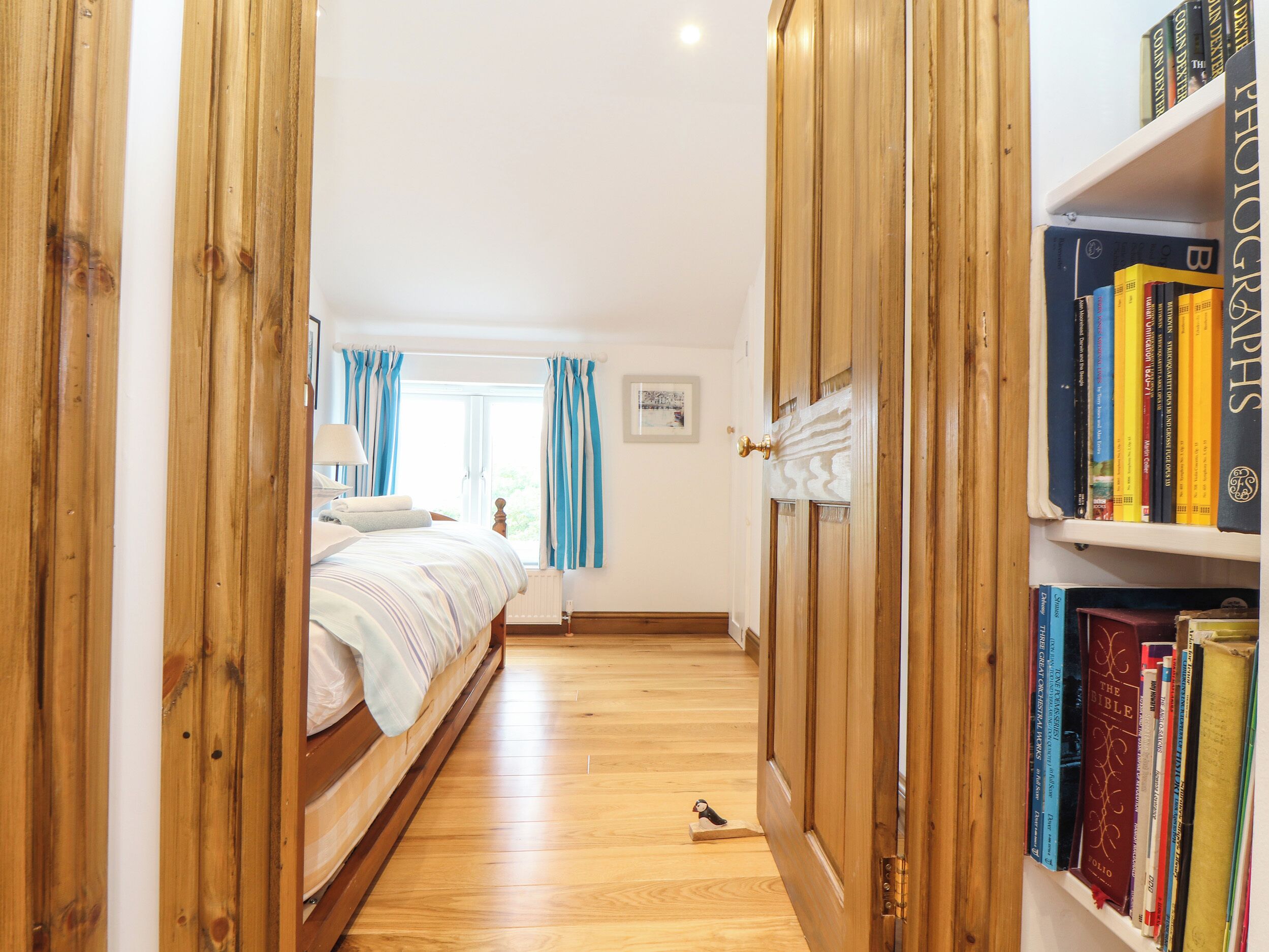 Un dormitorio con suelo de madera, una cama con ropa de cama blanca, una estantería llena de libros y una ventana con cortinas.