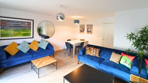 Een moderne woonkamer met twee blauwe banken, een houten salontafel, een eettafel met stoelen, een spiegel en een ingelijst sportfoto aan de muur.