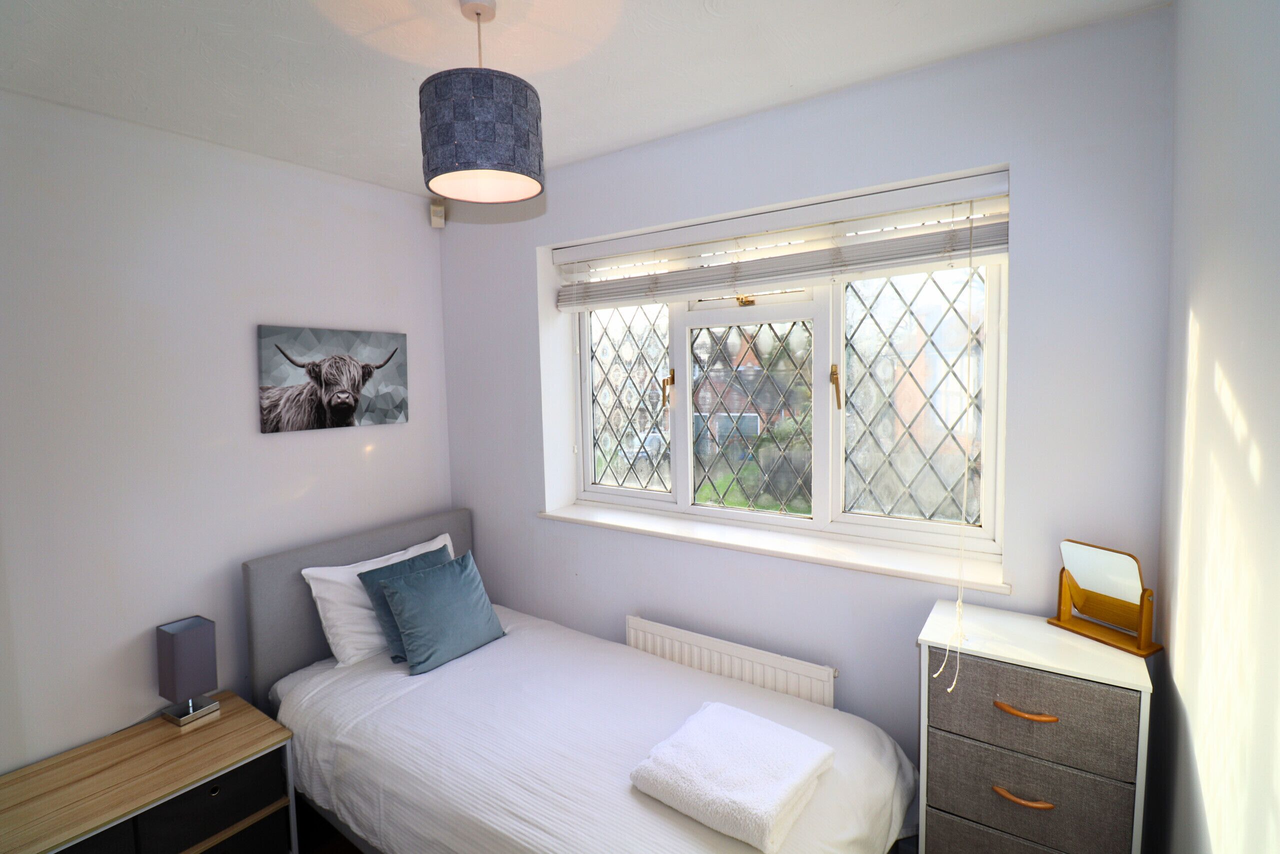 A bedroom with a bed, a bedside table, a dresser, a window with a view, and a hanging light fixture.