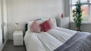 A bedroom with a bed, white nightstands, a bedside lamp, a potted plant, and a window with blinds. - Consett Shotley Bridge - Stylish Spacious Three Bedroom Apartment (Consett)