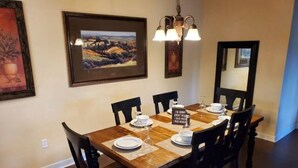 A dining room with a wooden table set for six, surrounded by six chairs. There is a chandelier above the table, and framed artwork on the walls.