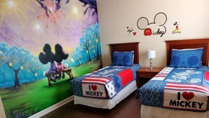 A room with two beds, each bedspread featuring a Mickey Mouse design and the text "I love Mickey". There is a wall mural of Mickey and Minnie Mouse in a park setting.
