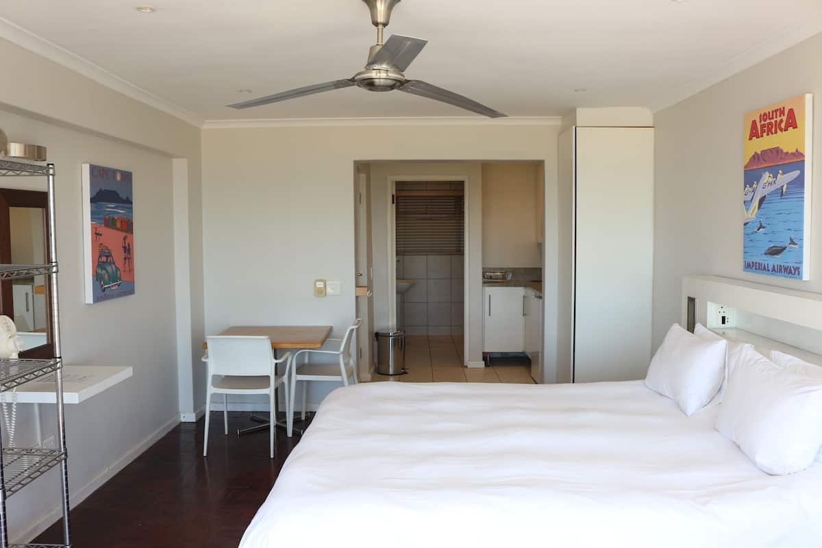 Une chambre d’hôtel avec un lit, un bureau, une chaise, un ventilateur de plafond, une salle de bain et une affiche au mur.