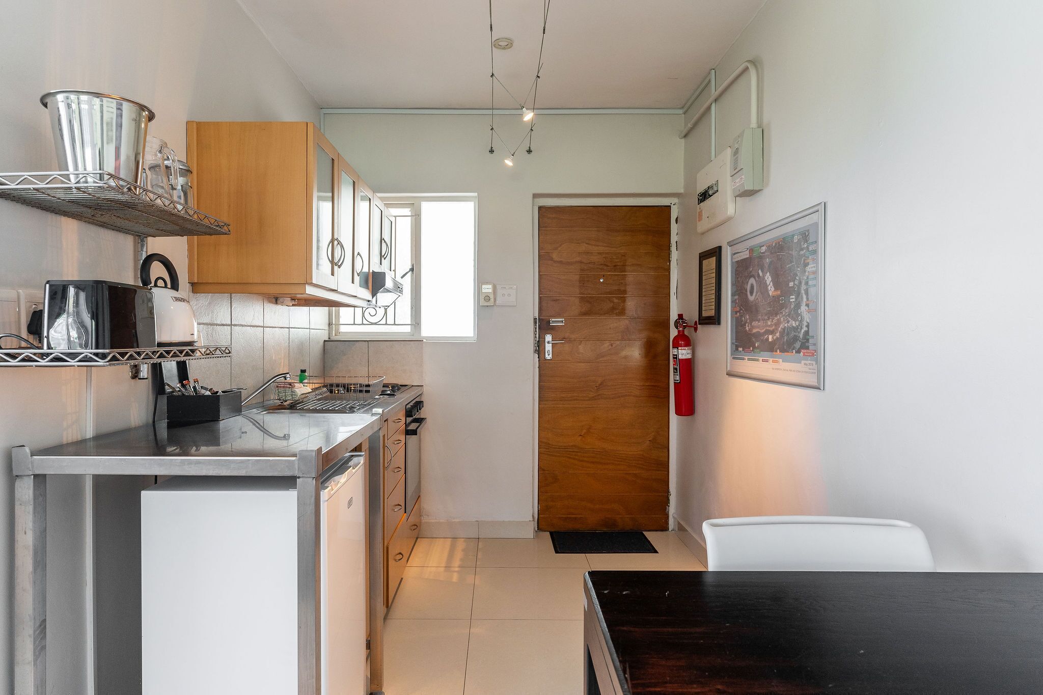 A compact kitchen with a refrigerator, microwave, toaster, and wooden cabinets. There is a wooden door leading to another room and a fire extinguisher mounted on the wall.