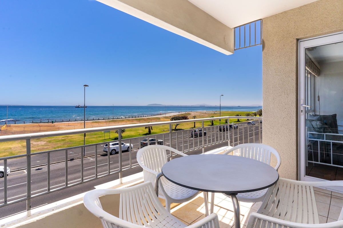 Un balcon avec des chaises blanches et une table, donnant sur une route et une plage, sous un ciel dégagé.