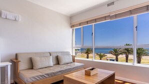 Een moderne woonkamer met een beige bank, een houten salontafel en grote ramen met uitzicht op het strand en de oceaan.