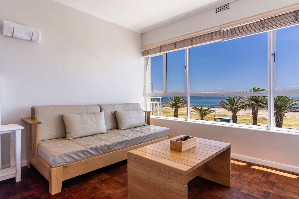 Un salon moderne avec un canapé beige, une table basse en bois et de grandes fenêtres donnant sur la plage et l’océan.