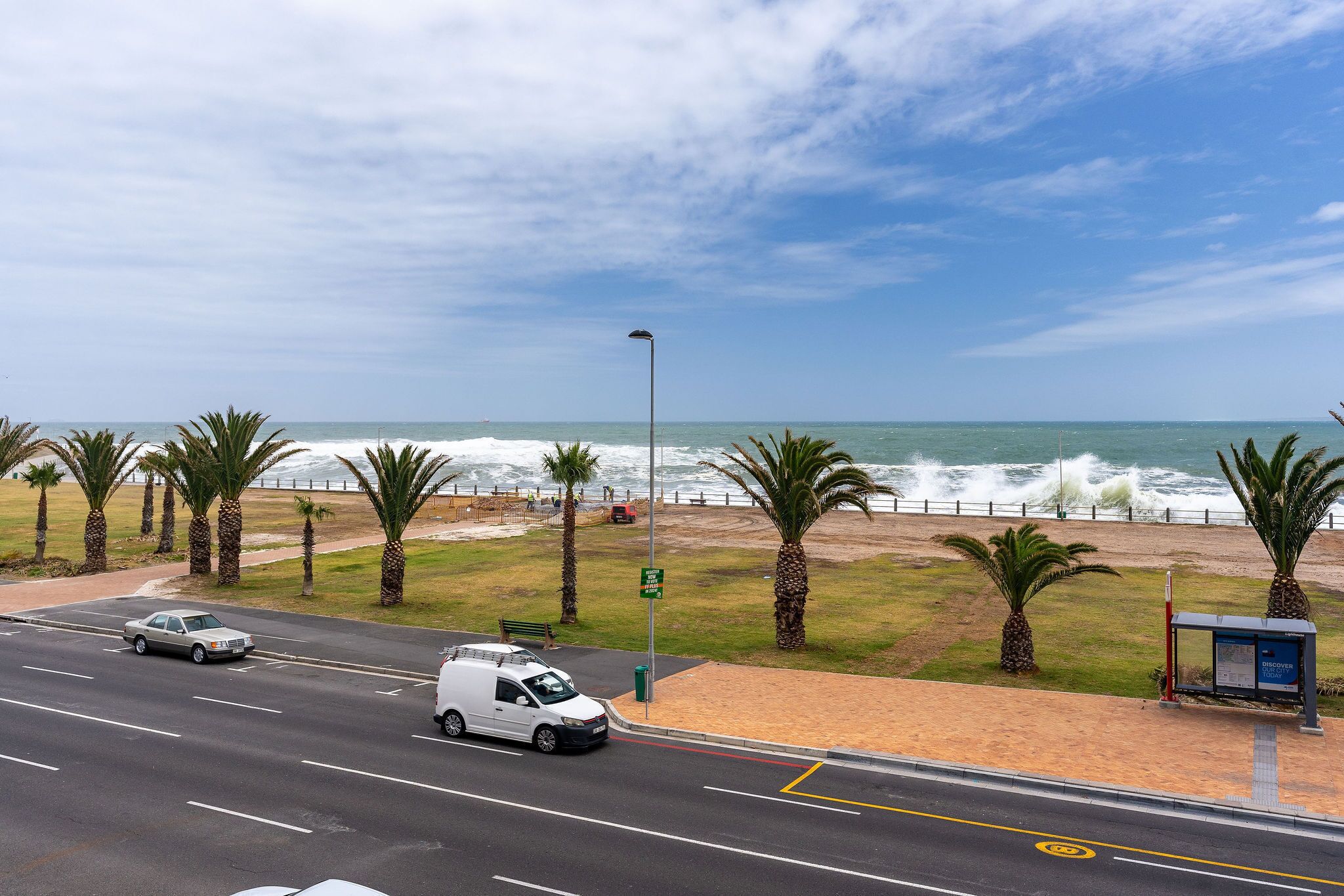 Carretera costera con vista a la playa, palmeras y una parada de autobús.