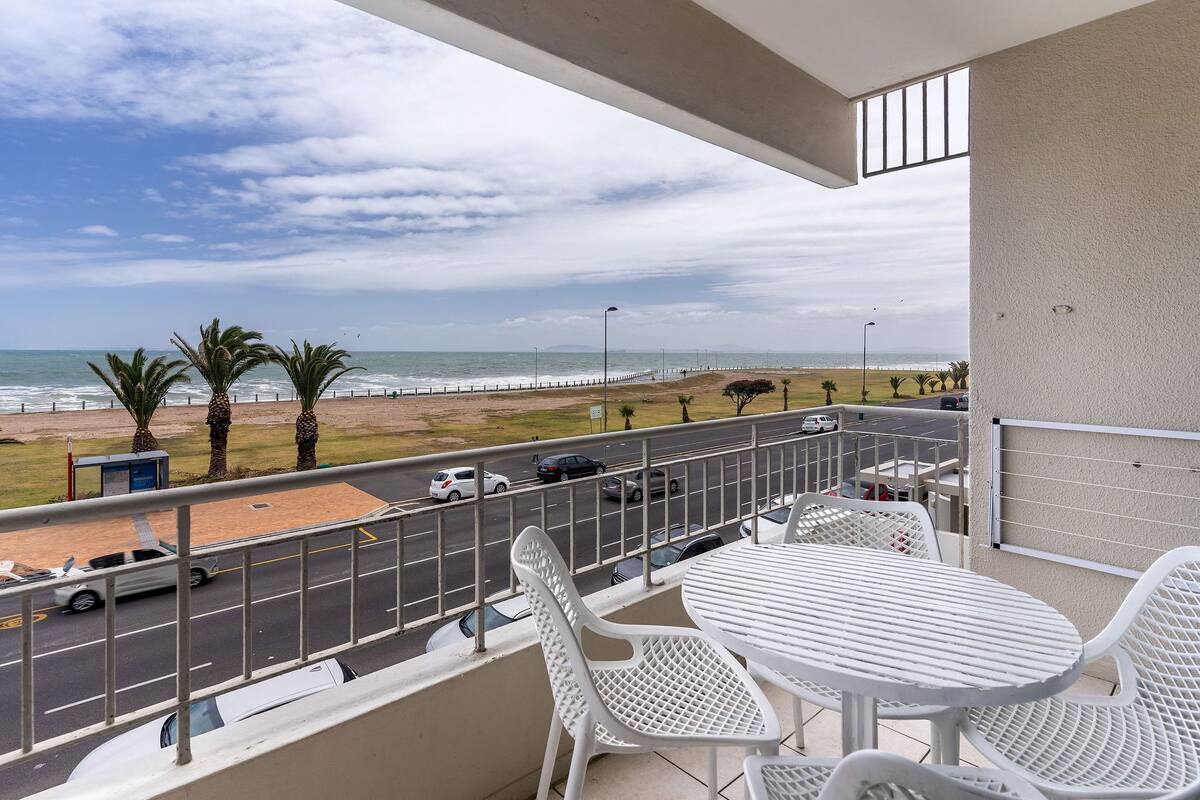 Un balcon avec des chaises et une table en plastique blanc, donnant sur une plage et une route où circulent des voitures.