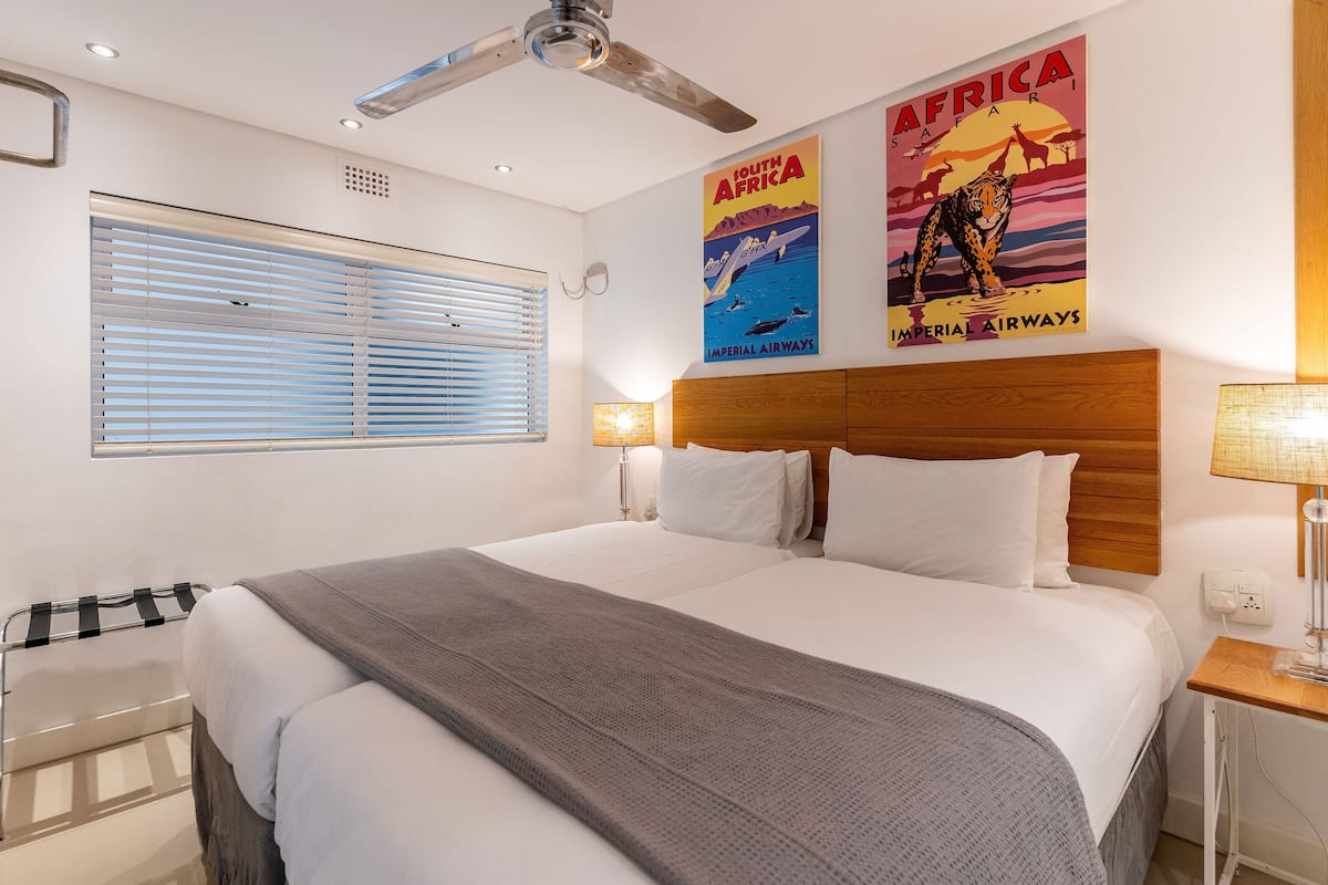Une chambre d’hôtel avec un grand lit, un ventilateur de plafond, une fenêtre avec des stores et deux affiches au mur.
