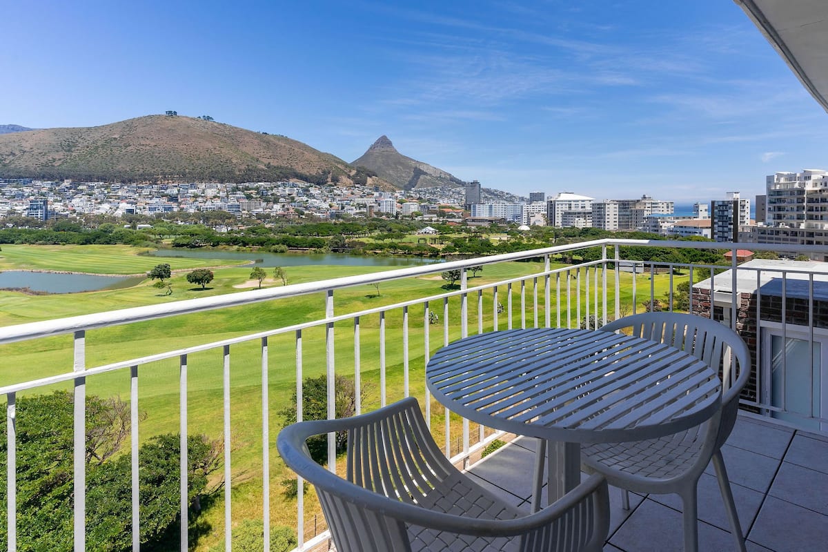 Un balcon avec une table et des chaises donnant sur un terrain de golf, une montagne et le paysage urbain.