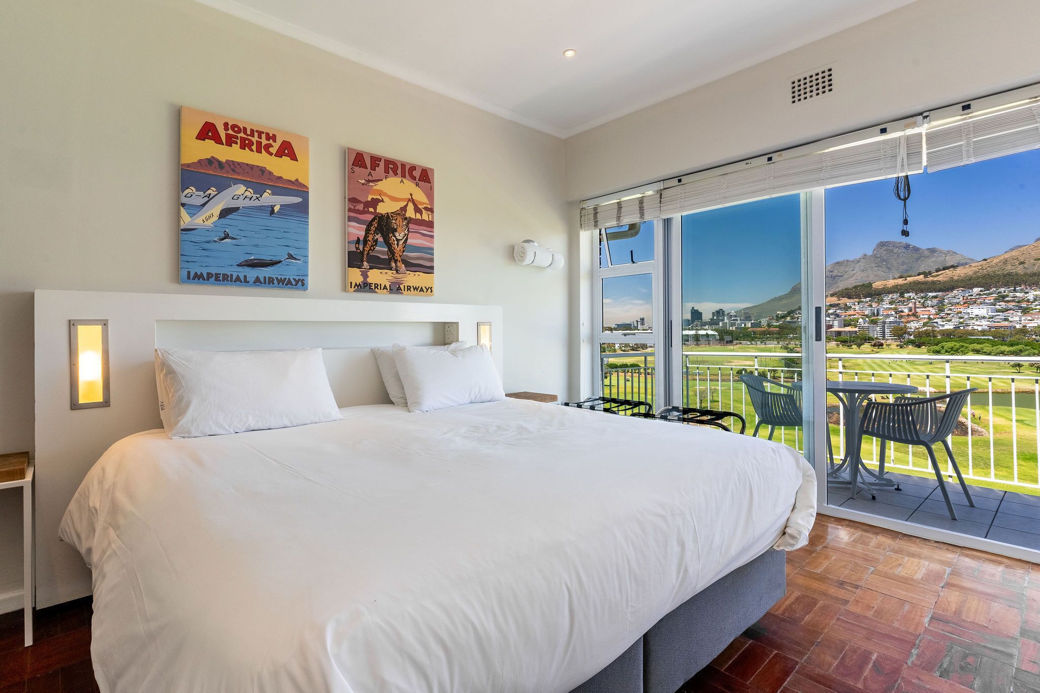 A hotel room with a large bed, a balcony with a table and chairs, and two posters on the wall.
