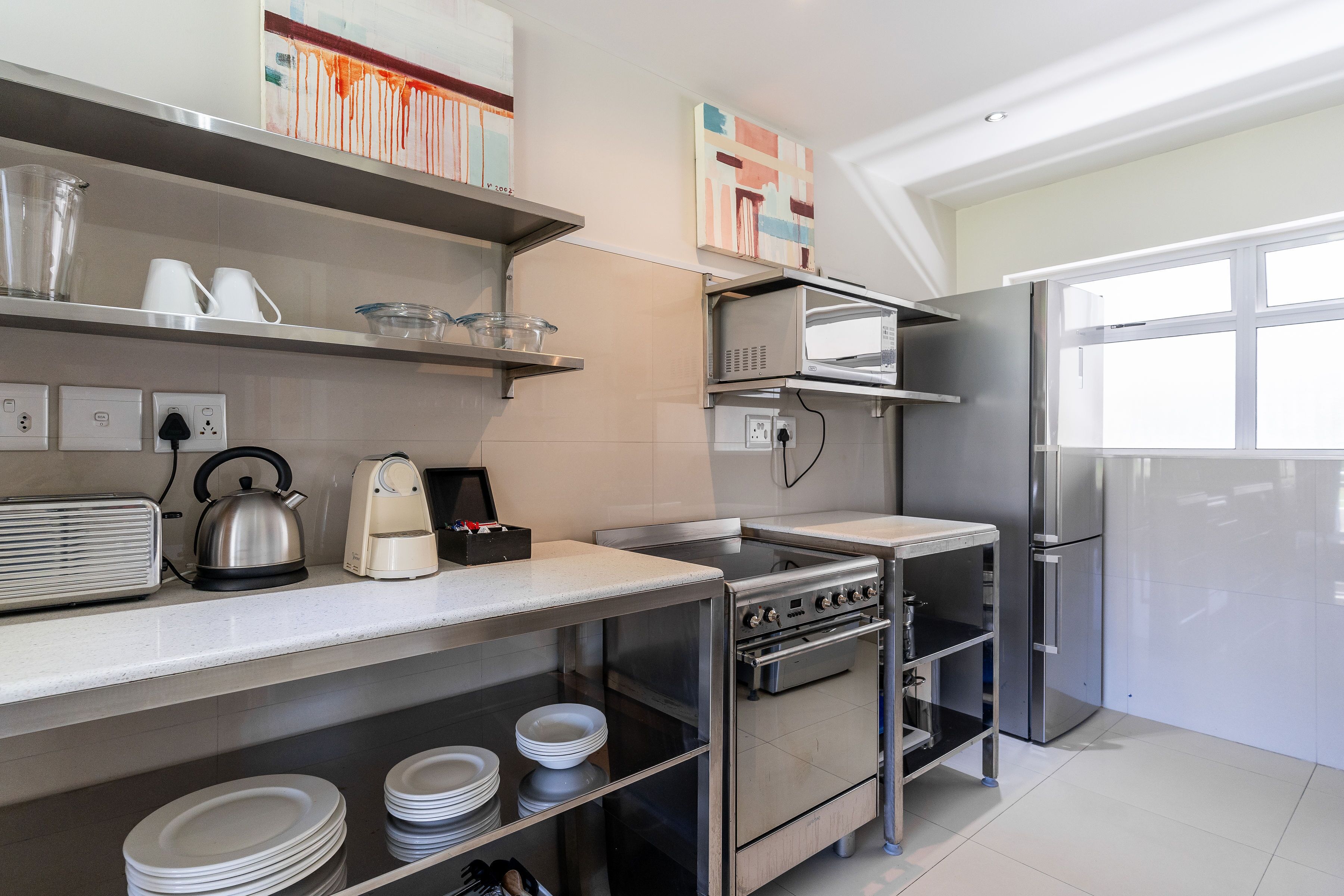 Una cocina moderna con electrodomésticos de acero inoxidable, incluyendo estufa, horno, microondas y refrigerador. Hay estantes sobre la encimera con vajilla y una tetera.
