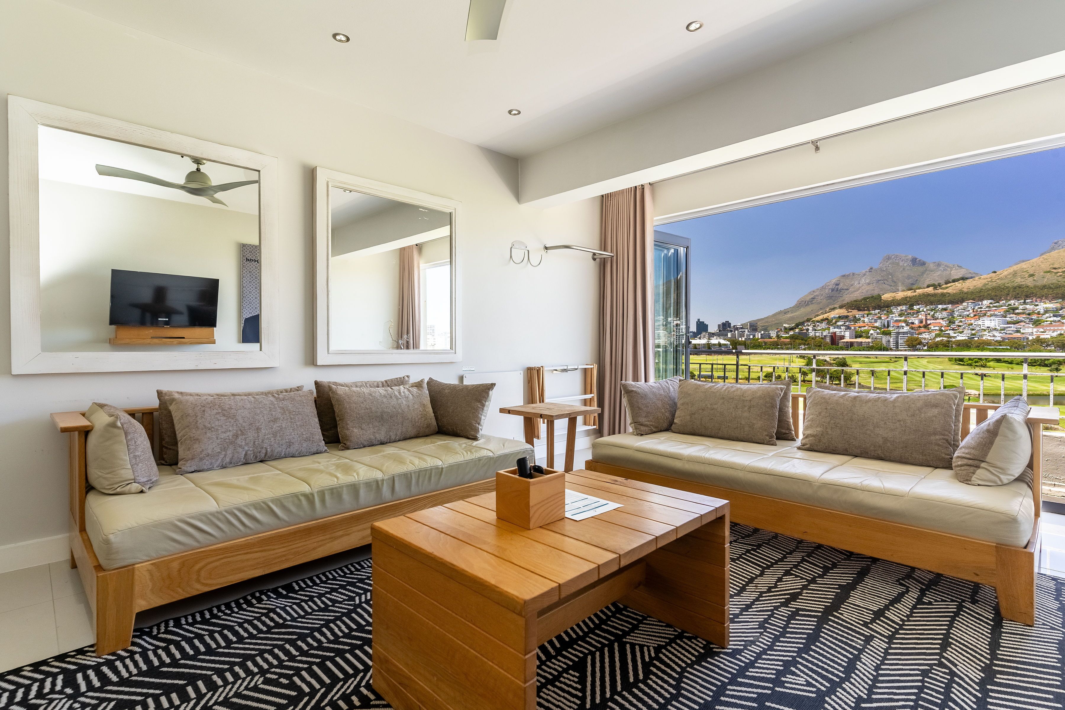 Una sala de estar moderna con dos sofás, una mesa de centro y un televisor. Amplias ventanas ofrecen vistas a la ciudad y a las montañas.