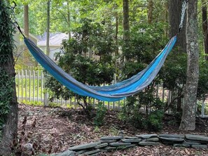 A hammock suspended between trees in a wooded area with a white fence in the background.