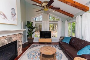 A living room with a stone fireplace, a ceiling fan, a large window with curtains, a brown leather sofa, and a wooden coffee table.