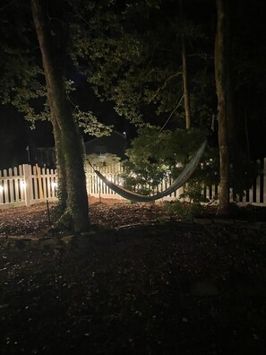 A hammock in a wooded area with a white picket fence in the background.