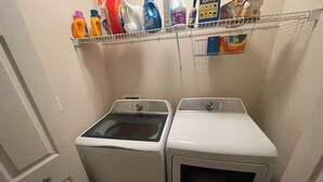 Un lavadero con dos lavadoras de carga frontal y un estante superior con varios productos de limpieza.