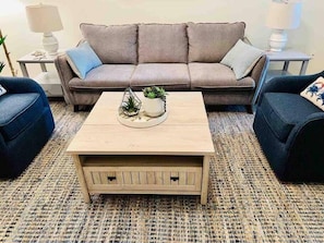 Ein Wohnzimmer mit einem beigefarbenen Couchtisch, zwei blauen Sessel, einem braunen Sofa mit Kissen und einem kleinen Beistelltisch mit einer Lampe.