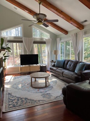 A living room with a television, sofa, and coffee table, large windows with curtains, and a ceiling fan. - Tranquil Waters, OceanPines, 3 br, Dog Friendly (Ocean Pines)
