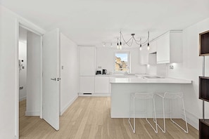 Une cuisine moderne avec des meubles blancs, un îlot central doté de tabourets de bar et une vue sur les bâtiments par la fenêtre.