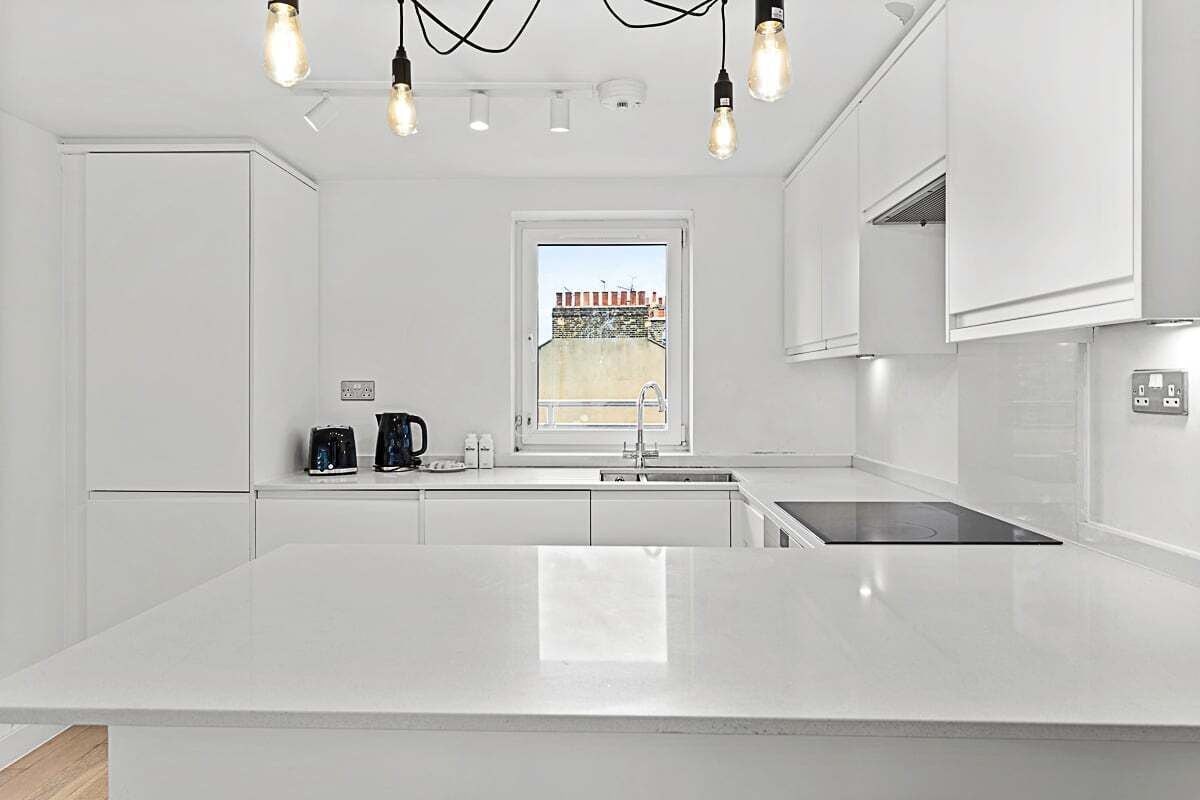 Une cuisine moderne avec des meubles blancs, un îlot central, une cuisinière intégrée et une fenêtre donnant sur des immeubles.