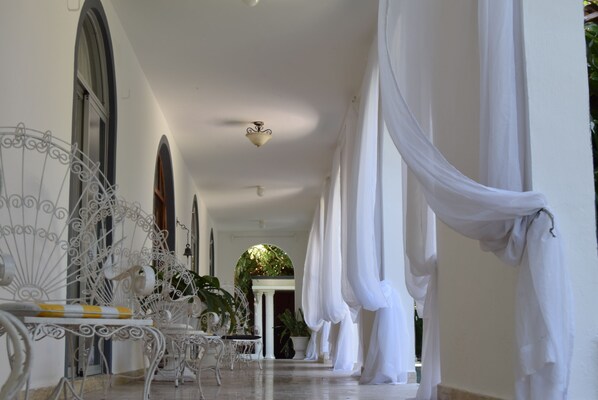 A corridor with white curtains, a white ceiling fan, and a white chair with a yellow and white striped cushion.