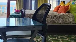 A room with a glass table, a chair, a sofa with colorful cushions, and a view of the sea through large windows.