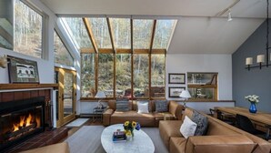 A spacious living room with a fireplace, leather sofas, a coffee table with flowers, and large windows offering a view of trees. - Vail Ski Home next to National Forest (Vail)