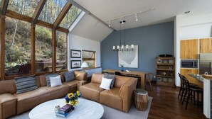 A modern living room with a large window, a sofa, a coffee table, and a kitchen area with wooden cabinets and a dining table. - Vail Ski Home next to National Forest (Vail)