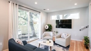 A modern living room with a sofa, armchair, and coffee table. There is a TV mounted on the wall, a large window, and a plant. - NEW Hip & Stylish House 30 steps from South Congress
 (Austin)