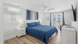 Ein Schlafzimmer mit Bett, Nachttischen, einem Schreibtisch, einer Klimaanlage, einem Deckenventilator und Blick durch eine Tür.