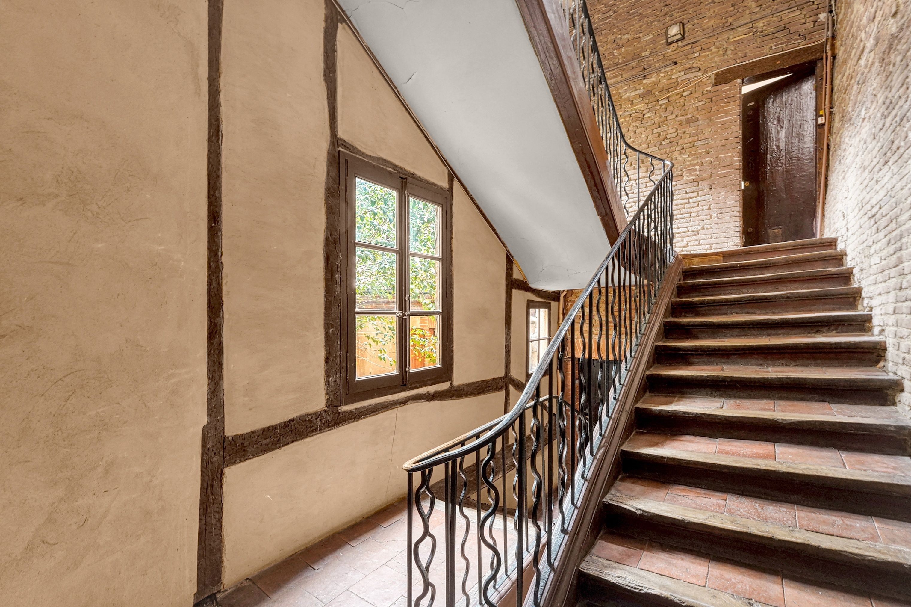 Un escalier avec une rampe en métal menant à une porte, une fenêtre donnant sur un paysage verdoyant et un mur dont la brique est apparente.