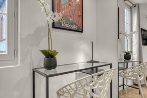 Ein modernes Zimmer mit Glastisch, weißen Stühlen, einer Topfpflanze und einem gerahmten Bild an der Wand.