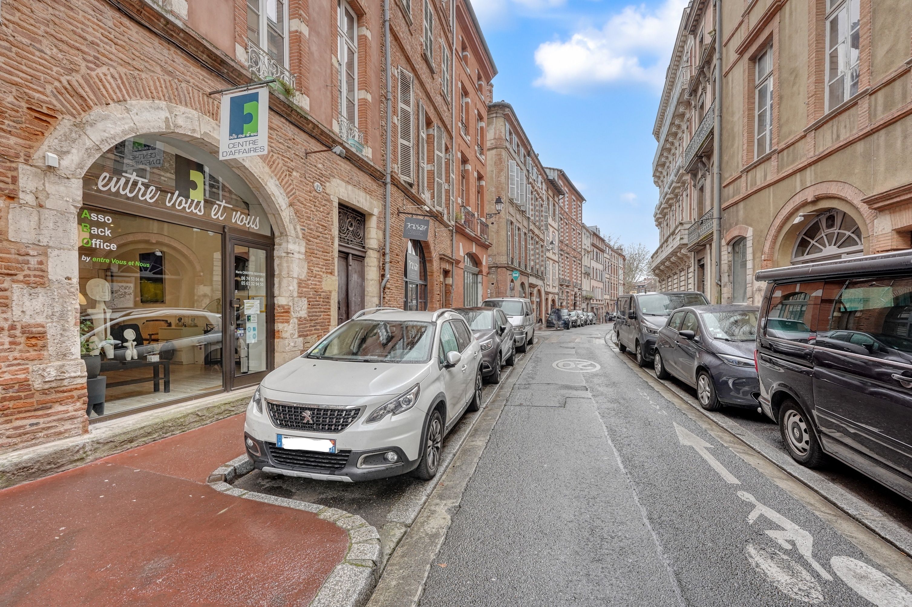 Une rue bordée de voitures garées et d’immeubles avec des enseignes.
