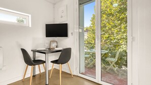 Ein kleiner Raum mit Tisch und zwei Stühlen, einem flachbildfernseher an der Wand und einer Glastür zum Außenbalkon mit Tisch und Stühlen.